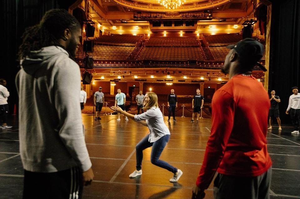 Lainie Hodges leading a session with a rookie class on a theater stage, players standing in a wide circle around her.